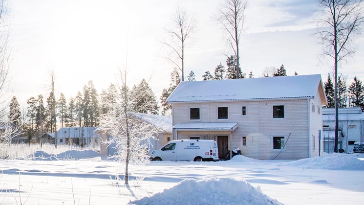 Vita och grå hus i vinterlandskap i Avesta. Vid huset som håller på att byggas närmast i bild kan vi se en arbetsbil utanför. Små björkar är täckta med snö bland husen.